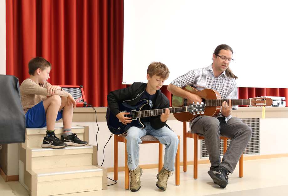 Ein Junge und ein Mann sitzen auf einer Bühne und spielen Gitarre. Ein weiterer Junge sitzt daneben und guckt zu.