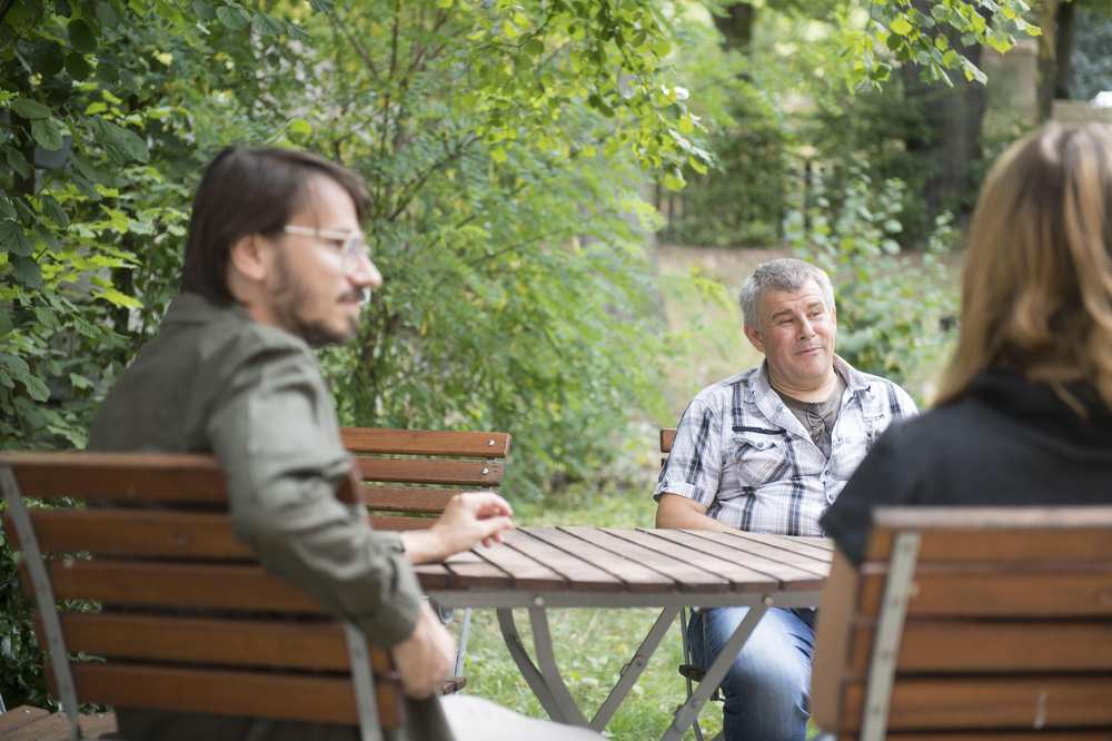 Drei Personen sitzen im Garten an einem runden Holztisch.