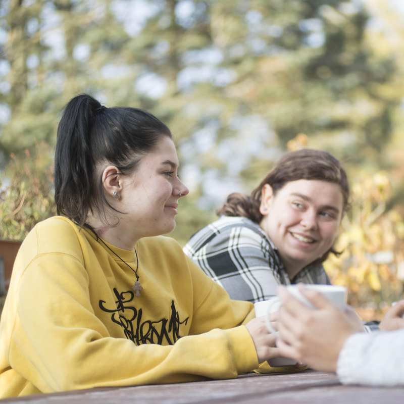 Drei Jugendliche sitzen an einem Tisch im Garten. Sie halten Kaffeebecher in den Händen.