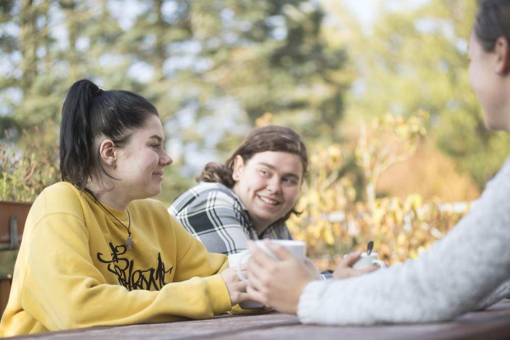 Drei junge Menschen sitzen am Tisch im Garten. Sie haben Kaffeebecher in der Hand