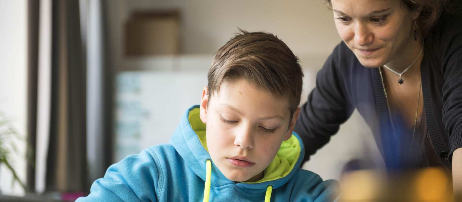 Ein Junge im Sweatshirt schreibt in ein Heft. Eine Frau schaut ihm über die Schulter.