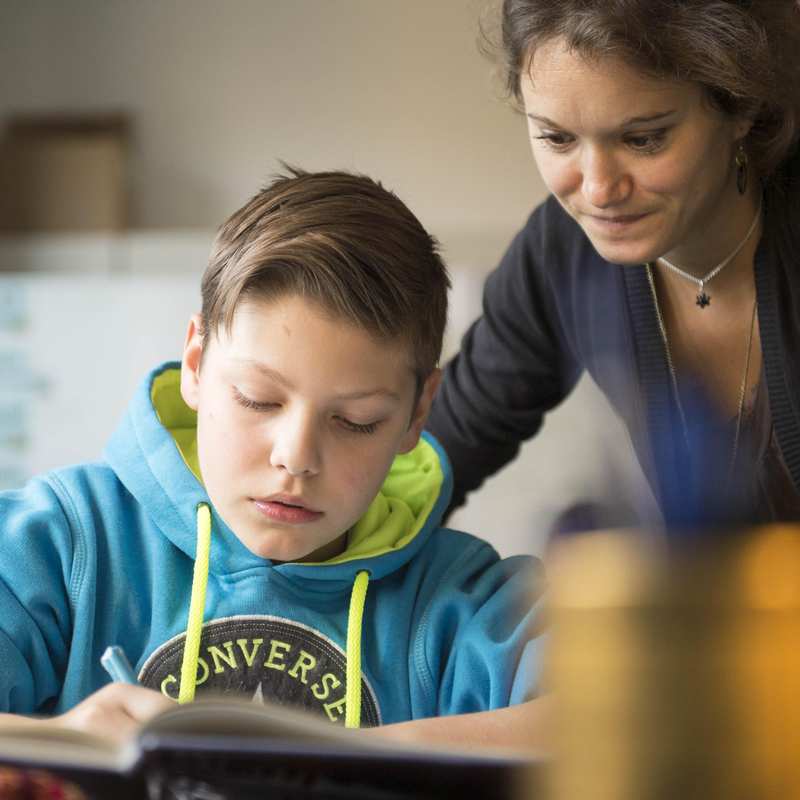 Ein Junge im Sweatshirt schreibt in ein Heft. Eine Frau schaut ihm über die Schulter.