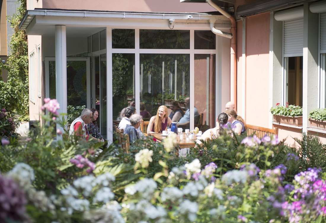 Eine Gruppe sitzt im sonnigen Garten vor einem Haus und unterhält sich.