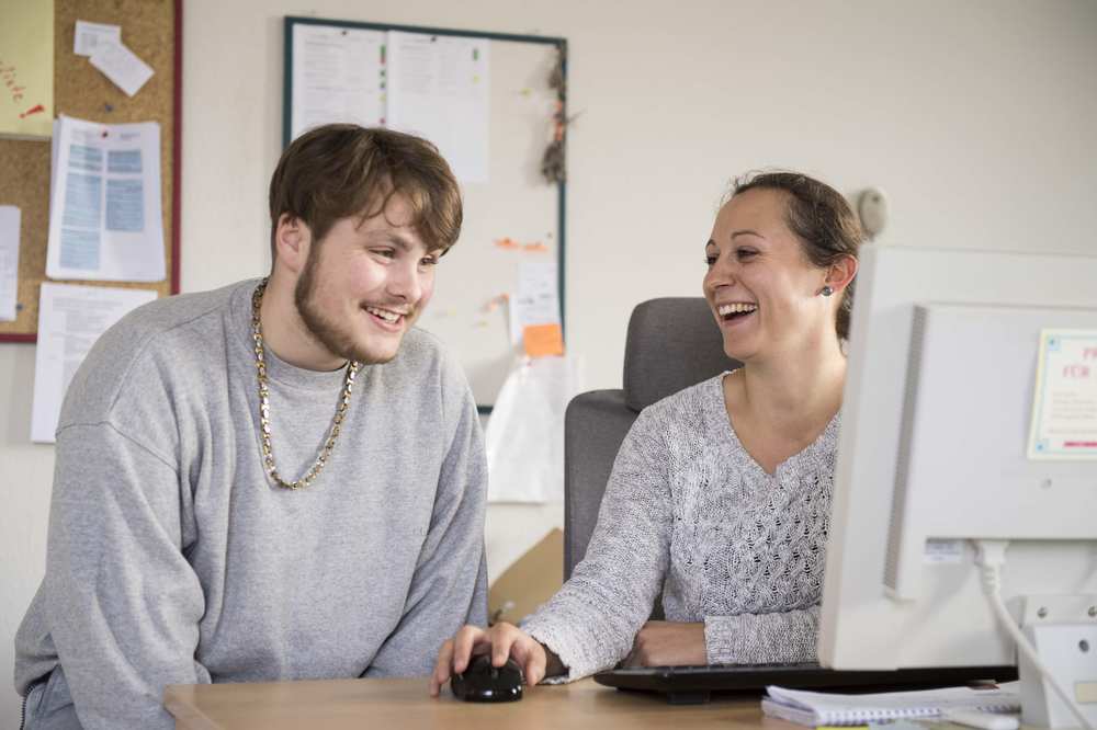 Eine junger Mann und eine junge Frau sitzen vor einem PC-Bildschirm und lachen.