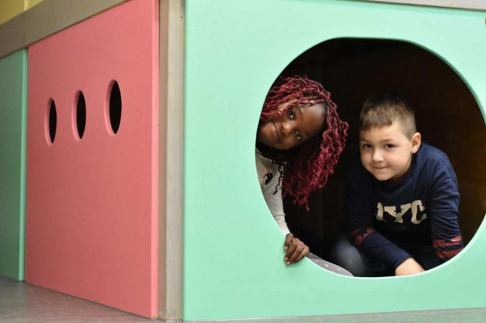 Zwei Kinder verstecken sich in einer Art Indoor-Höhle und schauen aus dem runden Loch.