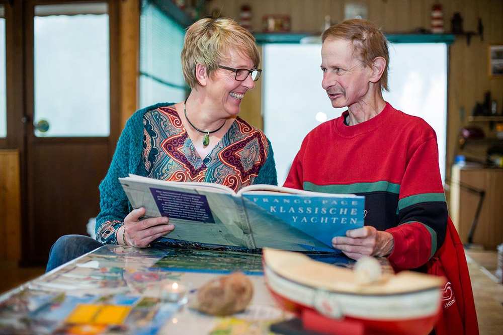 Eine FRau schaut sich gemeinsam mit einem kranken Mann ein Buch an. Sie sehen sich an.