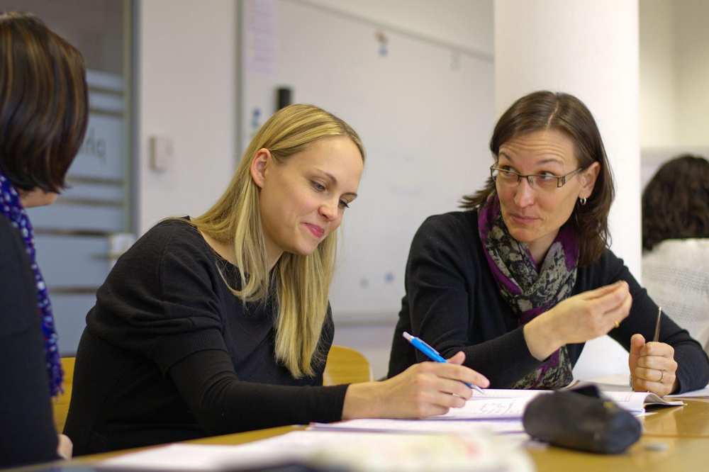 In der Mitte sitzen zwei Frauen am Tisch und sprechen miteinander.