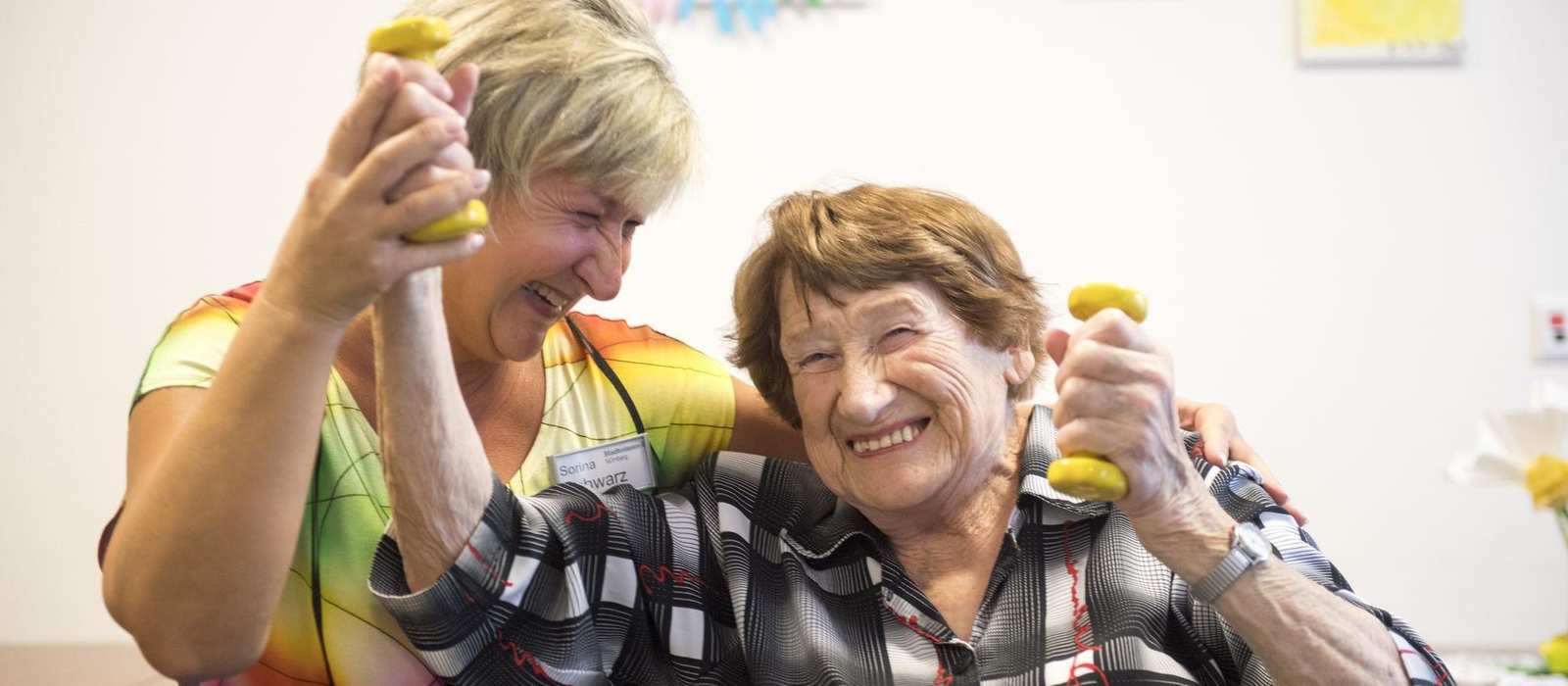 Eine Pflegerin macht mit einer Seniorin Gymnastik mit gelben Hanteln.