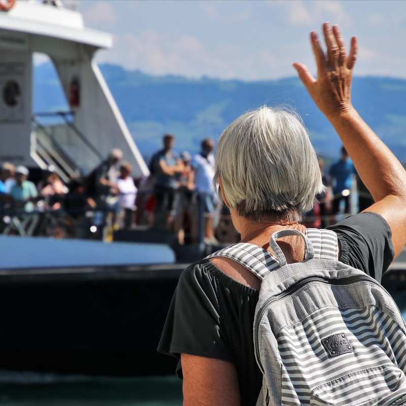 Das Bild zeigt eine Seniorin von hinten. Sie winkt Menschen auf einem Schiff zu.