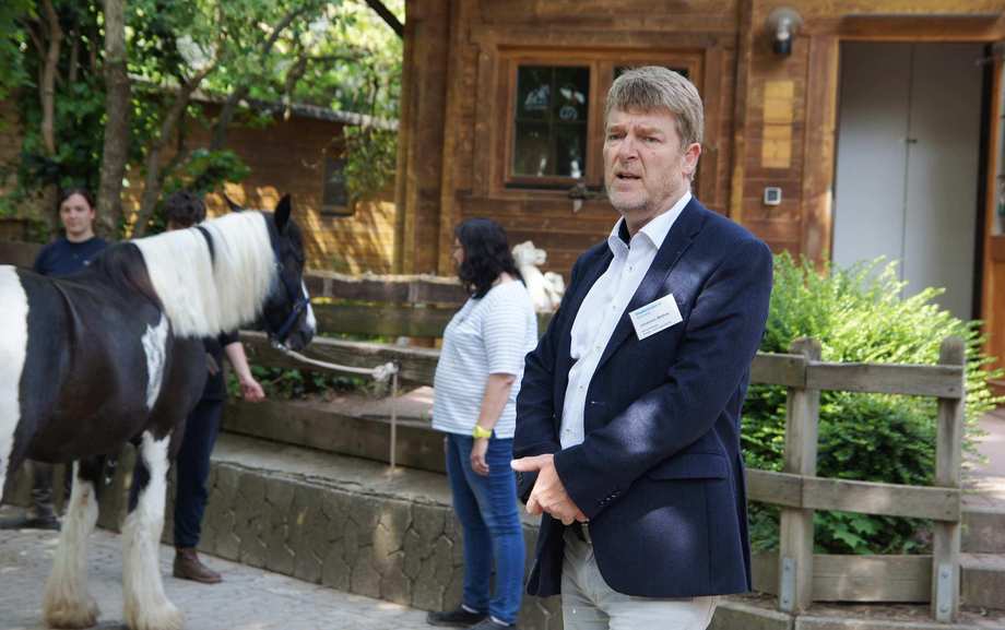 Ein Mann im weißen Hemd und blauen Jackett steht auf einem Hof. Im Hintergrund ein Pferd.