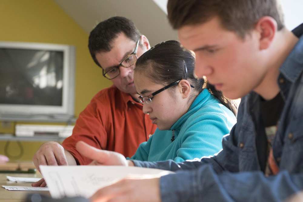 Zwei Schüler sehen in ihre Bücher. Ein Lehrer erklärt etwas.