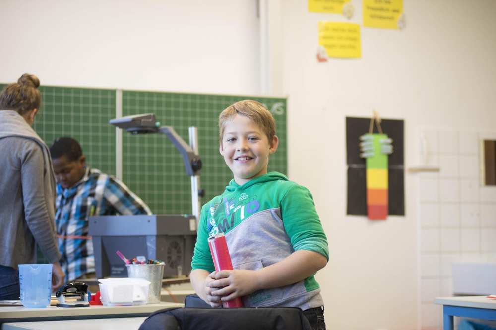 Ein Junge im  grünen Hoodie lacht. Im Hintergrund stehen ein Schüler und eine Lehrerin vor der Tafel.