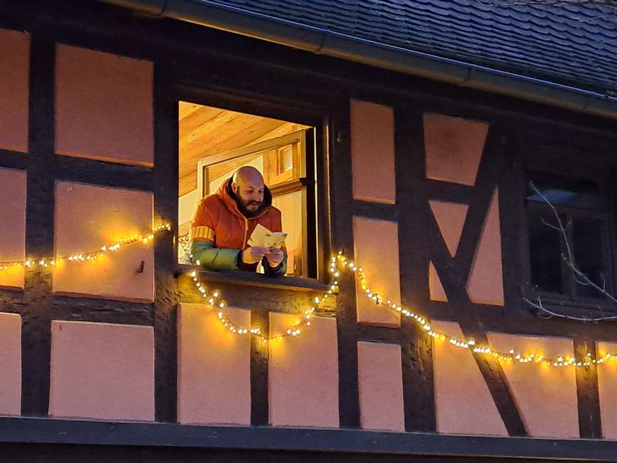 Mit lichtern geschmücktes Haus. Aus dem Fenster lehnt sich ein Mann und liest aus einem Buch vor.