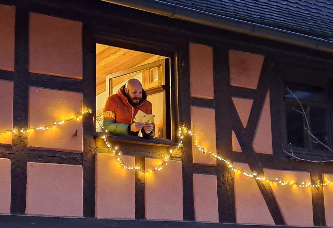 Mit lichtern geschmücktes Haus. Aus dem Fenster lehnt sich ein Mann und liest aus einem Buch vor.