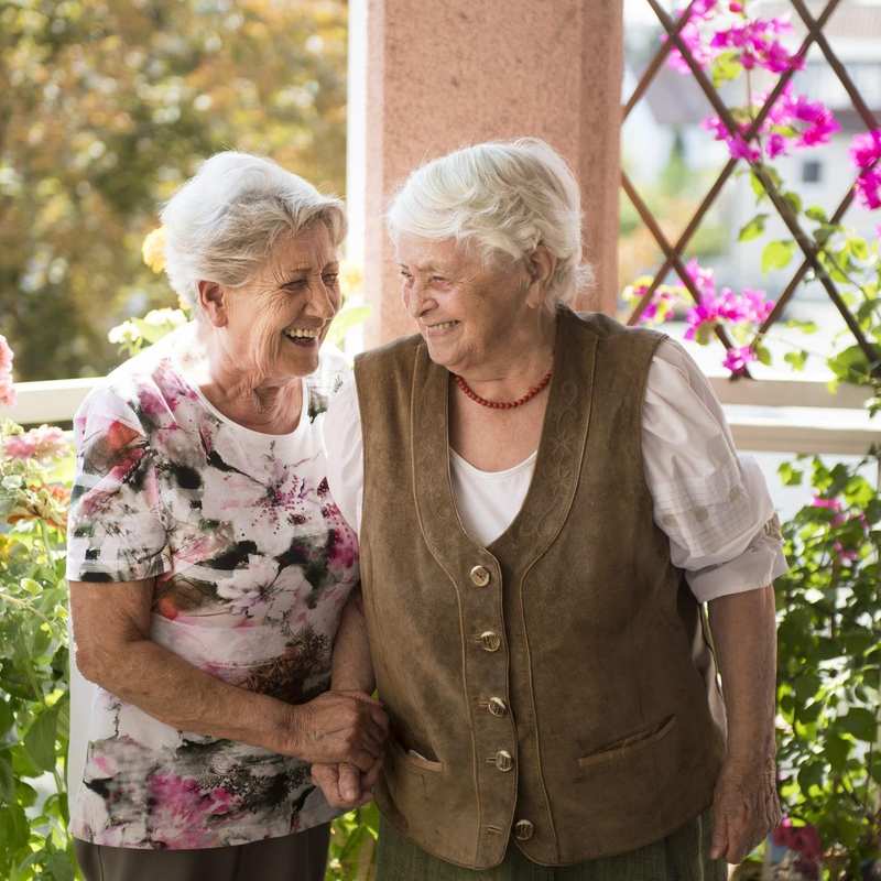 Zwei weißhaarige Frauen stehen lachend auf einer Terrasse.