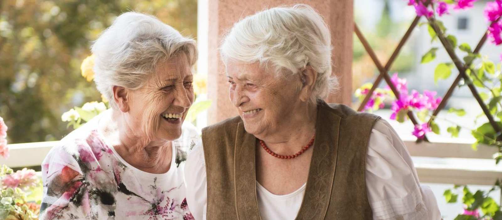 Zwei weißhaarige Frauen stehen lachend auf einer Terrasse.