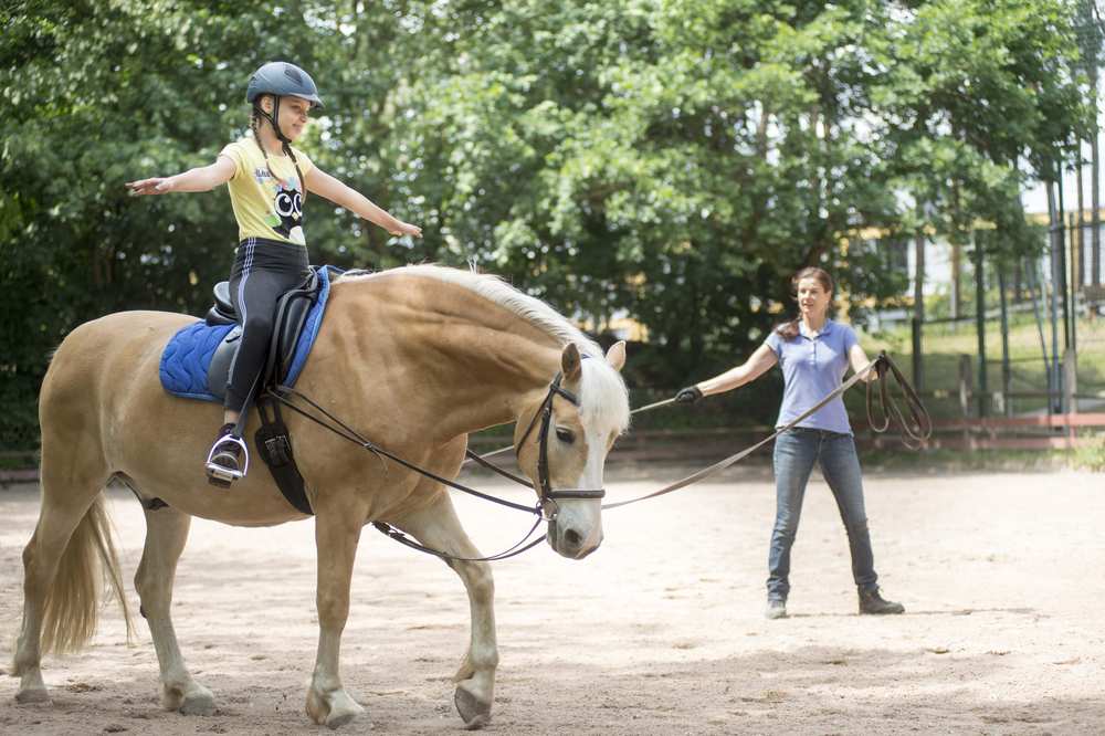 Ein Mädchen sitzt freihändig auf einem Pferd. Im Hintergrund steht die Reitlehrerin.